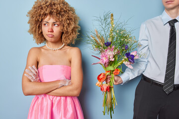 Offended curly haired young European lady keeps arms folded refuses to take bouquet from boyfriend turns aside wears pink dress and necklace isolated over blue background. Relationships problems