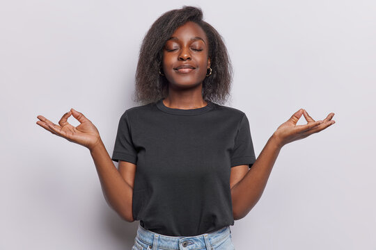 Calm and peaceful young African woman stands in lotus pose searches nirvana and patience with closed eyes wears casual black t shirt practices yoga or meditation isolated over white background