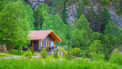 Fototapeta na wymiar House in forest. Cottage at foot of mountain. Villa in ecological location. Country house with terrace. Single storey wooden cottage. Scandinavian house in summer weather. Green trees surround villa