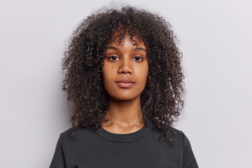 Portrait of serious curly haired teenage girl looks directly at camera has calm relaxed expression wears casual black t shirt isolated over white wall. Attractive female model poses for making photo