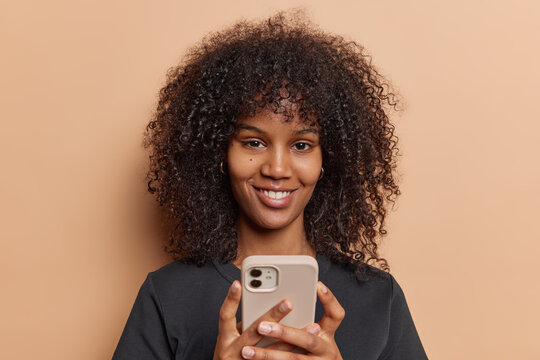 Portrait of lovely curly haired woman uses mobile phone for sending text messages enjoys online communication dressed in casual black t shirt isolated over brown background. Technology concept