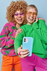 Gordijnen Vertical shot of overjoyed stylish women have fun make selfie via smartphone dressed in casual colorful clothes laughs at something funny stand next to each other isollated on blue studio wall © wayhome.studio
