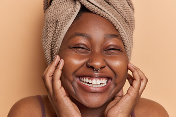 Close up portrait of dark skinned woman smiles toothily touches cheeks gently has soft clean skin after showering wears wrapped towel on head isolated over brown background takes care of herself