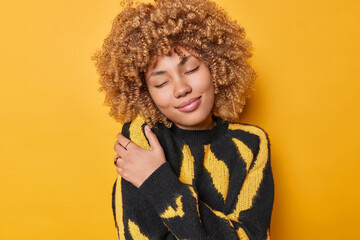 Lovely happy curly haired woman embraces herself daydreamsand embraces herself with both hands recalls romantic event wears comfortable jumper isolated over yellow background. Egoism concept - obrazy, fototapety, plakaty