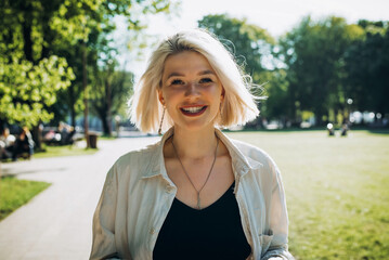 Naklejka premium Portrait of charismatic smiling blonde teenager in shirt walking in park on sunny day and looking at camera., lifestyle