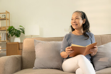 Asian woman in her 60s, happy retired woman reading a book on sofa in small living room. the comfort of her home