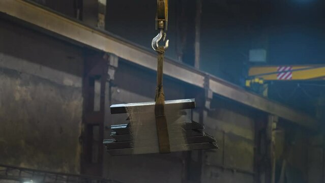 A load of metal pipes is moved on a overhead crane through a gloomy industrial space