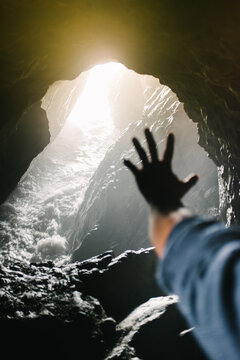 A man holds a silouetted hand out toward the entrance of a cave with water and sunlight streaming in