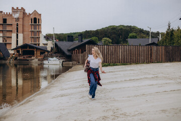 Charming beautiful girl in a white t-shirt and jeans holds a guitar in her hands, running along the lakeside, looking sideways and smiling. Positive cheerful woman is enjoying outdoor leisure.