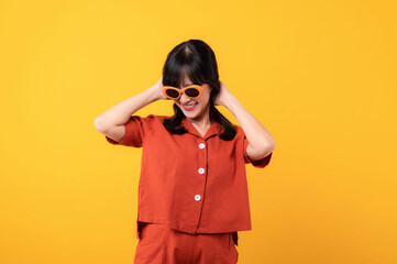 Portrait young beautiful asian woman cute and shy dressed in orange clothes and sunglasses touching cheeks showing shy emotional feeling isolated on yellow studio background.