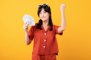 Portrait young beautiful asian woman happy smile dressed in orange clothes holding cash money and celebrate her success isolated on yellow background. Be rich concept.