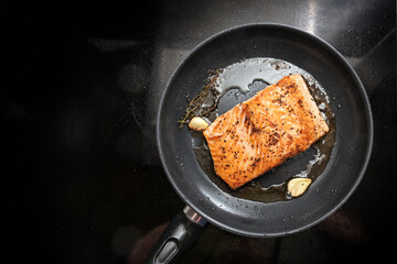 Fried salmon filet with with olive oil, herbs and garlic in a black frying pan on the stovetop, copy space, high angle view from above, selected focus