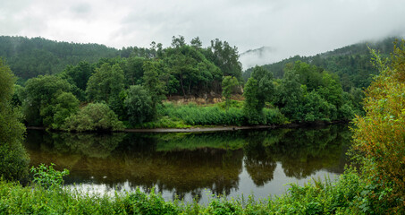 View of the green fluvial landscape reflected in the waters of the Confluence of the Miño river and the Avia river, Ribadavia Orense, summer of 2021, Spain