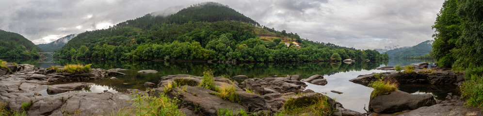 Panoramic landscape with a river, large stones in the foreground and a mountain with green trees in the background on a cloudy summer day of 2021 in Galicia, Spain.