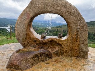 Fuente de piedra con forma de círculo en un paisaje de montañas verdes al fondo y nubes, verano de 2021 Galicia, España.