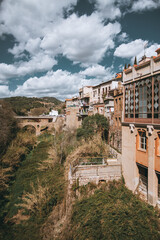 Fototapeta na wymiar Caldes de Montbui; a vertical shot of the blue sky with clouds. Lush vegetation is located behind the warm-colored buildings, adding a natural backdrop. A charming Roman bridge completes the scene