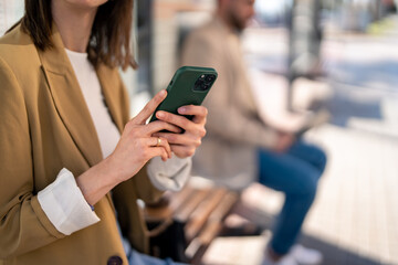 Hands of unrecognizable woman holding mobile phone while sitting at bus station and waiting for...
