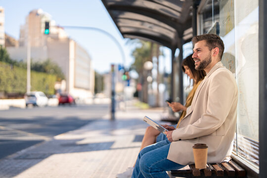 Side view of handsome young confident business millennial man with takeaway coffee sitting at bus station, holding digital tablet device waiting for bus in the city. Photo with copy space.