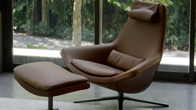 Comfortable brown leather armchair with footrest standing in modern living room with panoramic windows on background. Home interior concept