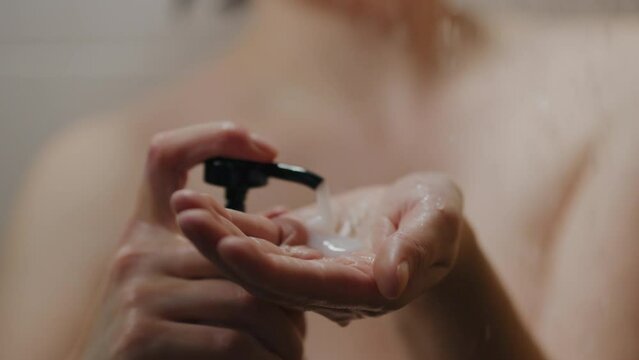 A girl of European appearance is washing herself in the shower, standing under a tropical shower, lathering her hands with shampoo, applying it to her hair Girl holding shampoo stands on falling water