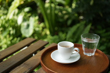 White coffee cup and glass of water on wooden table with green lush background