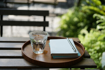 A glass of water and notebook on inside a wooden tray on wooden table