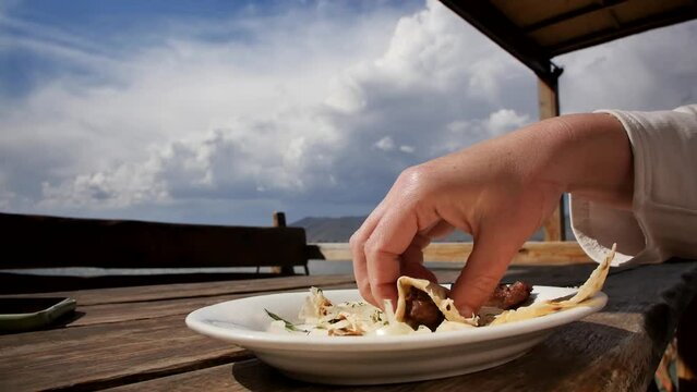 The hand of a white woman takes with her fingers a piece of oriental pita bread and fried meat on a plate during lunch at a restaurant table in the open air near the sea and mountains