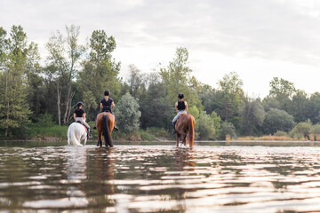 Three rider girls crossing the calm river water riding their beautiful horses on the cloudy summer...