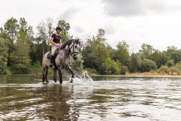 Girl riding gray horse down the calm river water with forest greenery reflections from the...