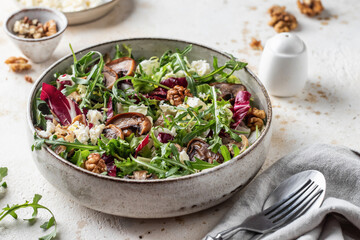Green salad with feta cheese, arugula, mushrooms, walnut in ceramic bowl on textured background. Healthy summer lunch or dinner
