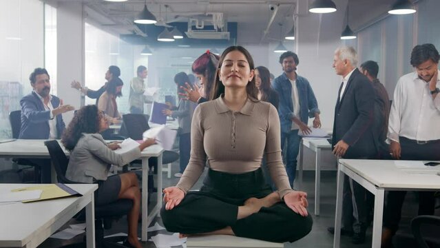 A young Asian Indian corporate female executive sitting quietly eyes closed in a meditative pose middle of a busy chaotic and messy office where people or coworkers throwing papers, fighting, shouting