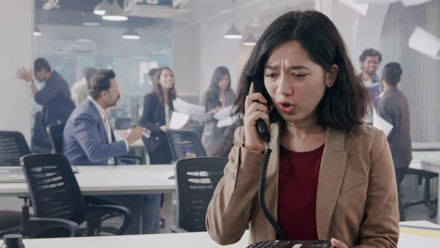 An angry stressed Asian female corporate office executive employee shouting or talking loudly on a landline phone in a chaotic start up business with people throwing papers and fighting in the back