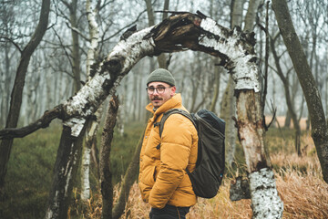 Portrait of young smiling friendly man with yellow winter jacket and hat, that looks behind his shoulder and stands under bending tree, during cold cloudy autumn day, Decin, Czech republic