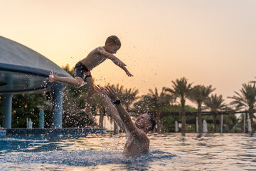 Dad and son are playing in the swimming pool. The father tosses the son up.