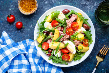 Nicoise salad with canned  tuna, tomatoes, boiled eggs, green beans, potatoes and olives on white plate, blue table background, top view