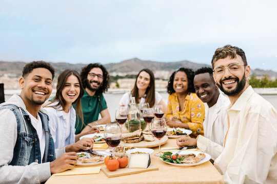 Group of friends gathered smiling at camera sitting together at a table enjoying barbecue dinner celebrating a party in summer. Portrait of multiracial people