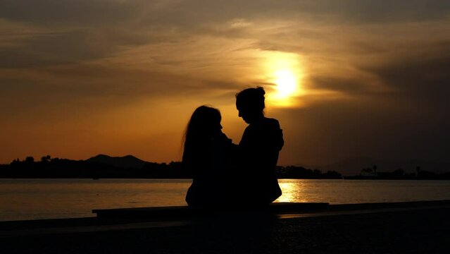 Silhouette of mother kiss child. A calm mother with her little daughter silhouettes sit on the river bank and pass the time during summer evening.
