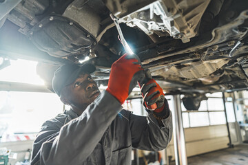Car mechanic with LED work light repairing vehicle chassis, standing under it. High quality photo