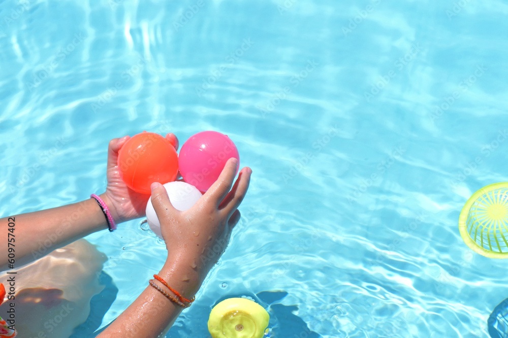 Wall mural Baby girl play toy in swimming pool on a hot summer day. Kids learn to swim. Child hold balls in water toys. Children play in tropical resorts. Family beach vacation.