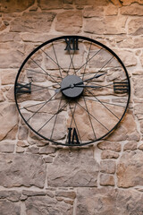 Black clock in the form of a bicycle wheel on the background of a stone wall. Thematic hours related to bicycles. The concept of punctuality