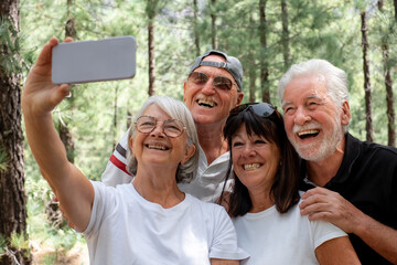 Four cheerful senior people in the mountain park looking at phone for a selfie - Group of smiling retiree enjoying nature and trekking day together