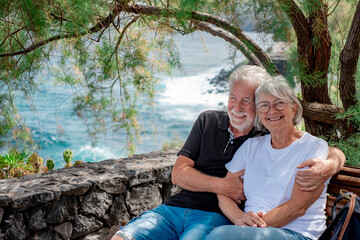 Lovely old senior family couple embracing, romantic elderly people sitting in outdoor on shade of a plant enjoying moment of affection cuddling. Sea on background