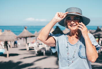Happy senior woman in hat sitting near beach talking on mobile phone enjoying freedom and beautiful sunny day, relaxed senior lady on vacation or retirement using smartphone