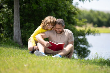 Lovely family reading a book on grass. Boy and dad having a picnic in park. Happy son and father reading a book. Happiness family. Father reading a book to child sitting outdoor on the grass in park.