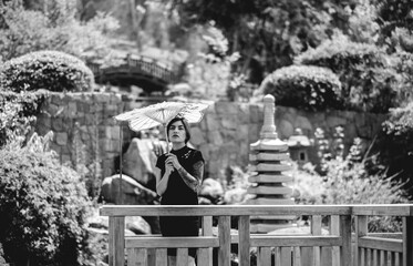 Fototapeta premium Captivating beauty: brunette with tattoos, chic dress, and paper umbrella, delighting in a sunny day at a japanese garden (in black and white)