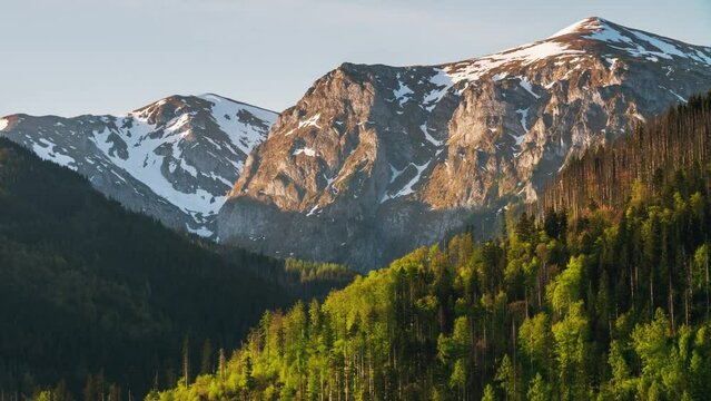Early morning sunrise over alpine mountains time-lapse shot, snow covered mountain range peaks and green hills with coniferous forest lit with warm morning sunlight, moving shadows, rising sun