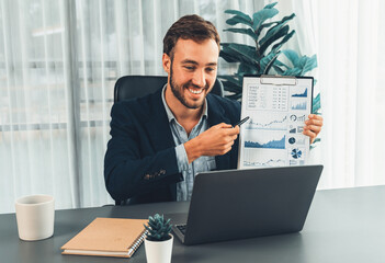 Professional businessman in black suit present financial data or BI paper clipboard to laptop camera during online presentation meeting. Remote work with virtual meeting presentation in office. Entity