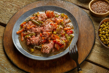 Israeli couscous in spices with sumac, chicken pieces and vegetables in a plate on a wooden round stand next to a fork.