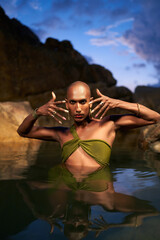Androgynous bipoc lgbtq model poses in water inside natural pool at night. Non-binary person shows...