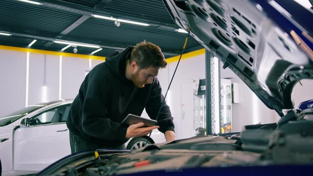 A male auto mechanic with a tablet in his hands stands at the open hood of a luxurious blue car that has broken down and repairs it